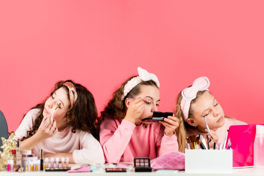 three young girls put on makeup in front of a pink backdrop.